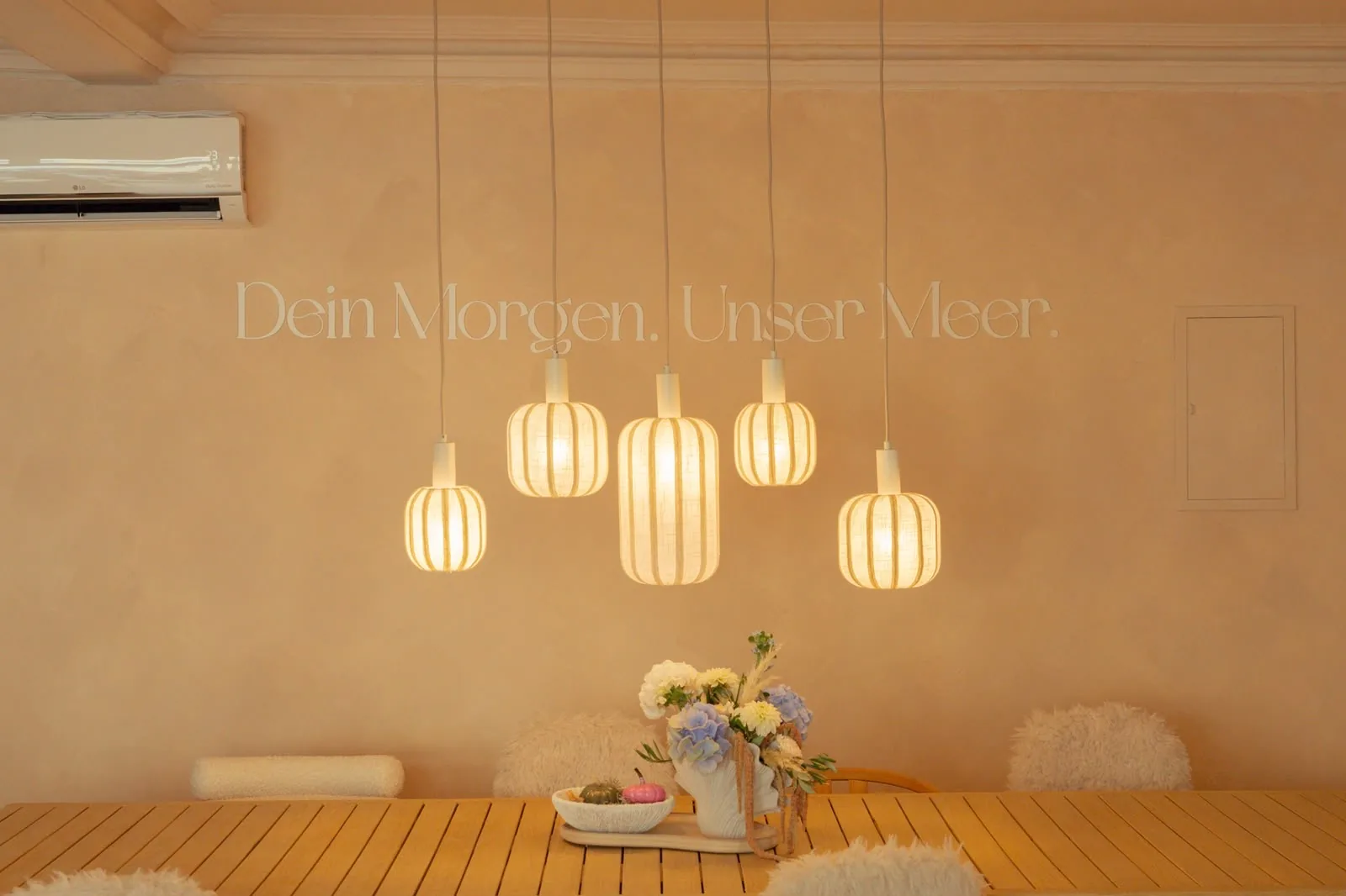 Heller Gastraum mit fünf warm leuchtenden Pendelleuchten vor einer Wand mit dem Schriftzug „Dein Morgen. Unser Meer.“, davor ein Holztisch mit Blumen.
